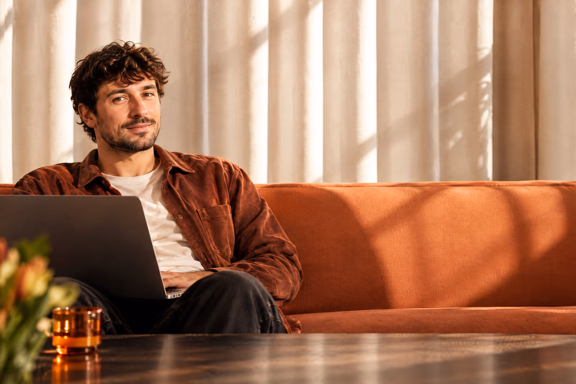 A student on a rust sofa with a laptop on his knees, smiling at the camera